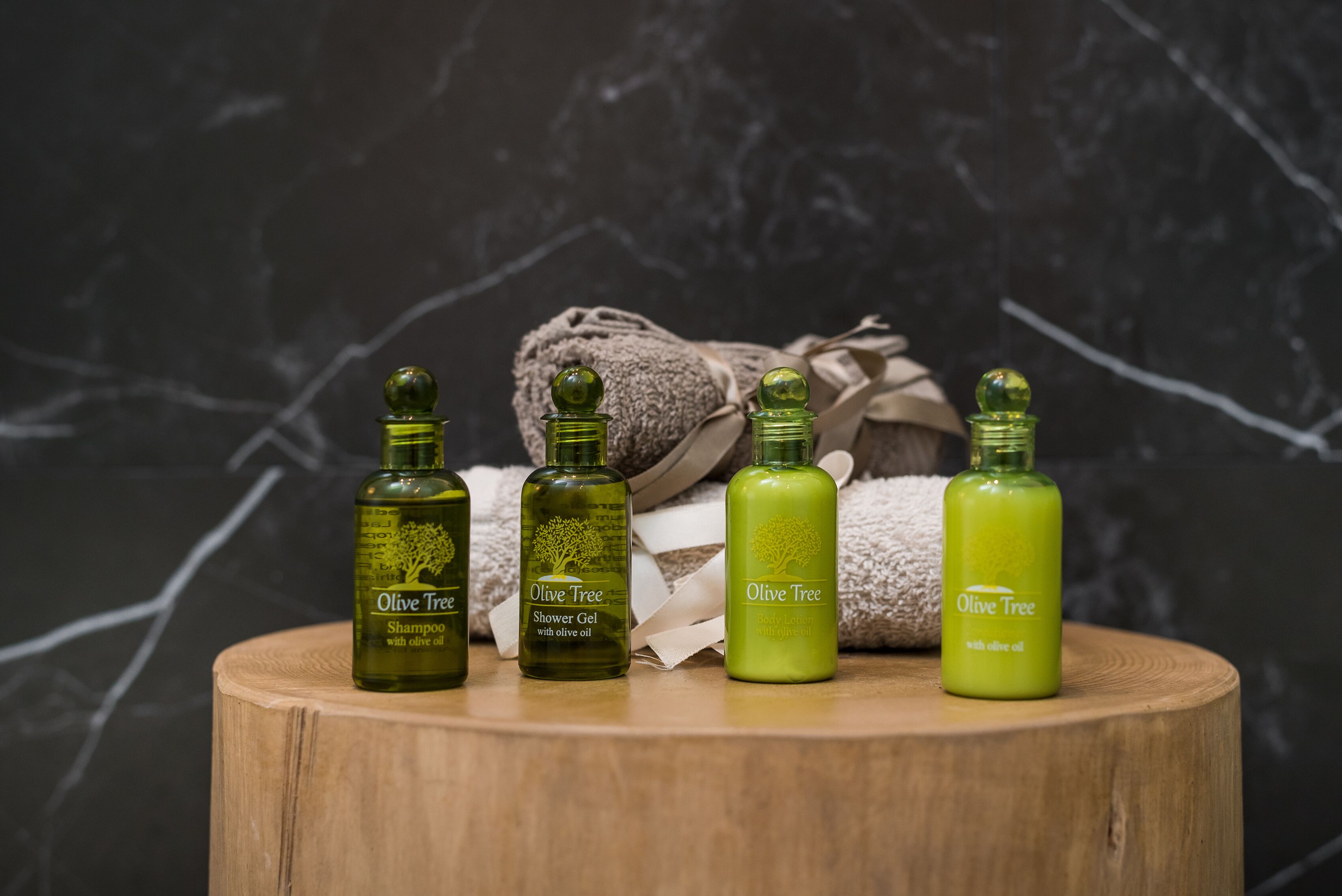 Four bottles of Olive Tree products on a wooden surface with a towel and a rolled-up towel in the background.
