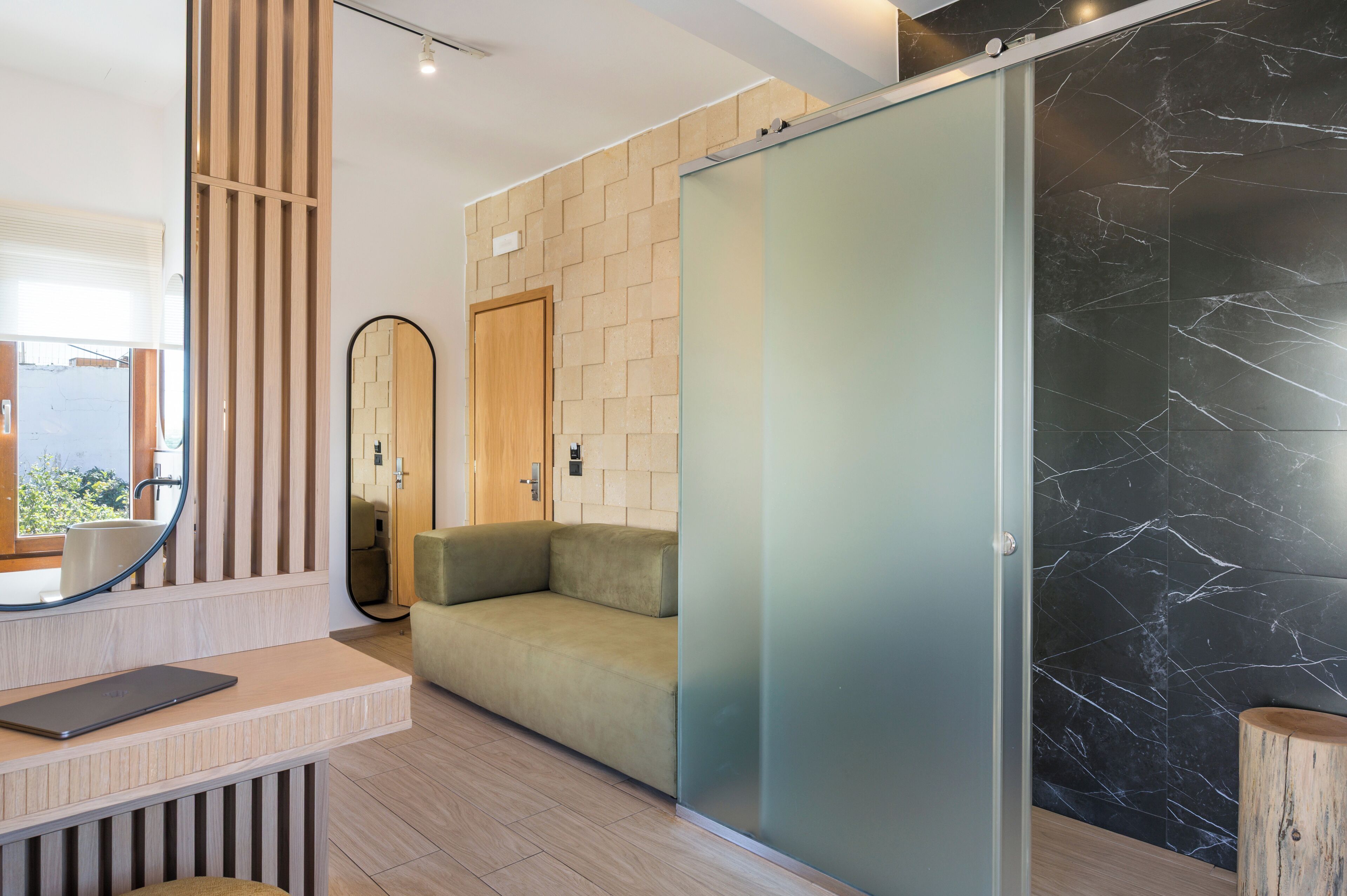 Quarto moderno com porta de vidro deslizante, sofá, escrivaninha de madeira com um laptop, espelho e parede com padrão semelhante ao mármore.