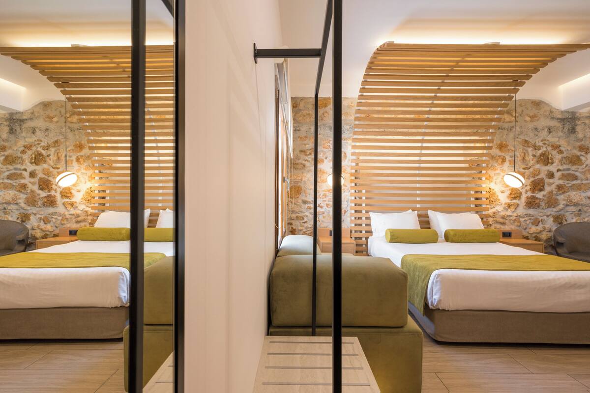Habitación de hotel con dos camas, cabeceros de madera con listones y paredes de piedra, con una lámpara moderna.