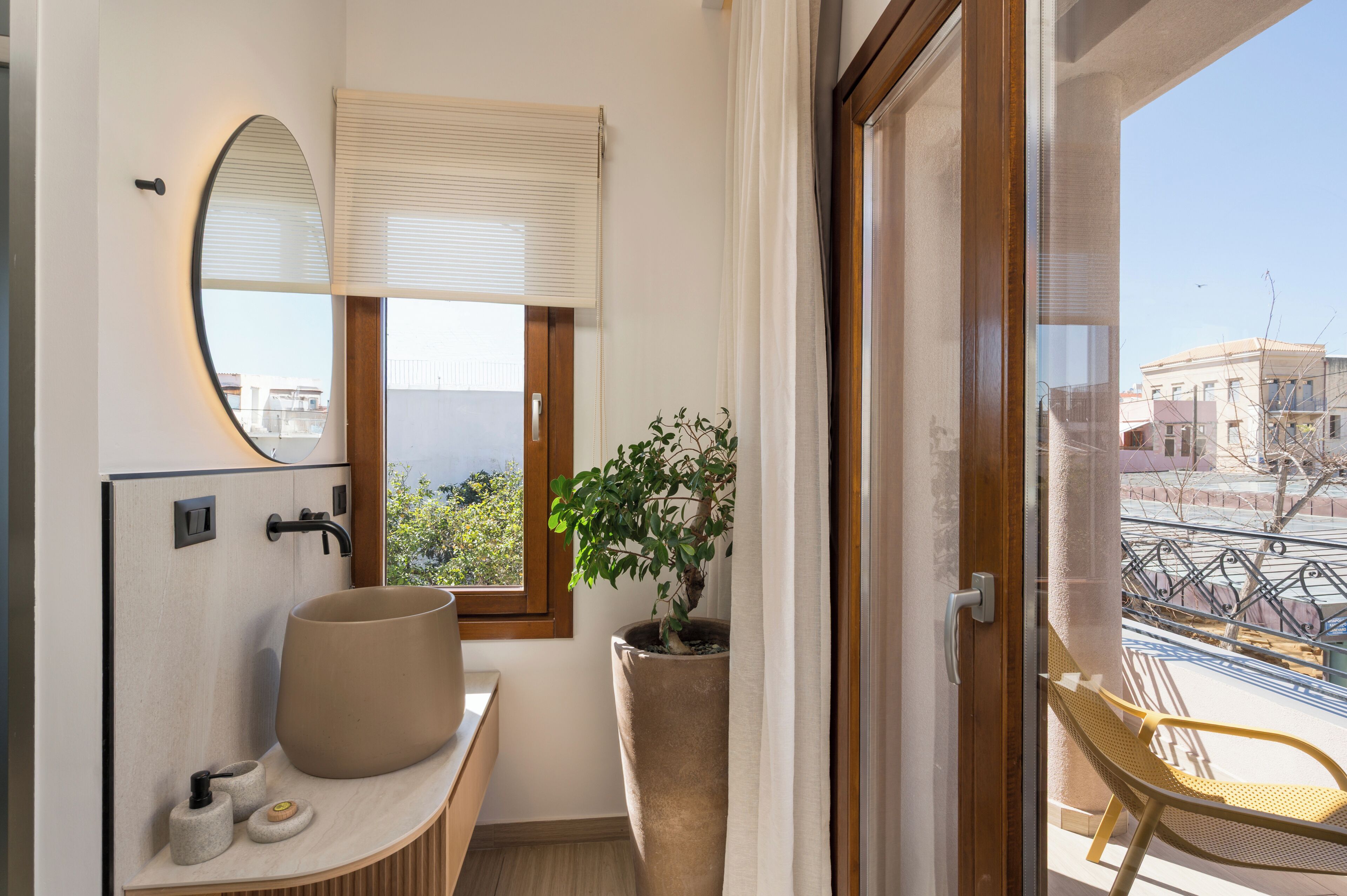 Un baño con un espejo ovalado grande, un lavabo con una pileta color beige, una planta en maceta y vistas a un balcón con una silla.