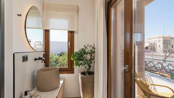 Un baño con un espejo ovalado grande, un lavabo con una pileta color beige, una planta en maceta y vistas a un balcón con una silla.