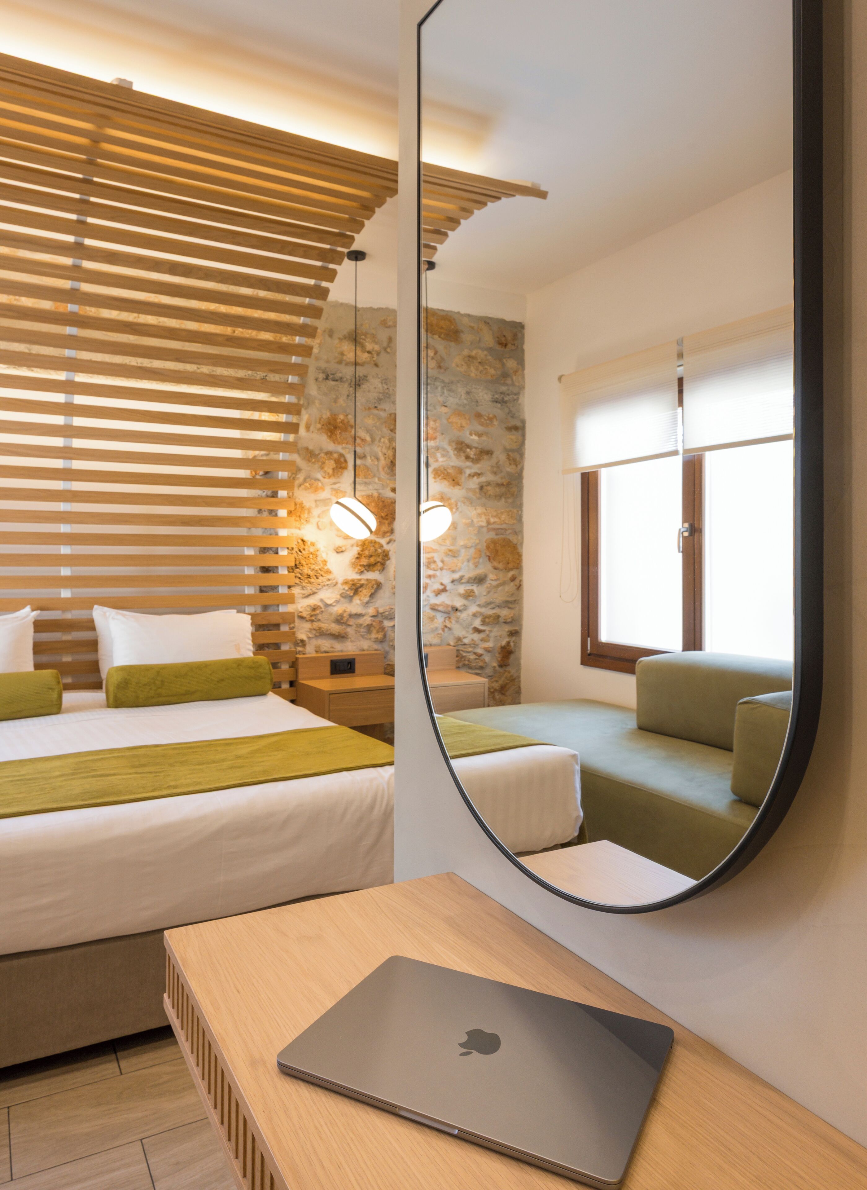 A hotel room with a bed, a sofa, a desk with a laptop, a mirror, and a stone wall.
