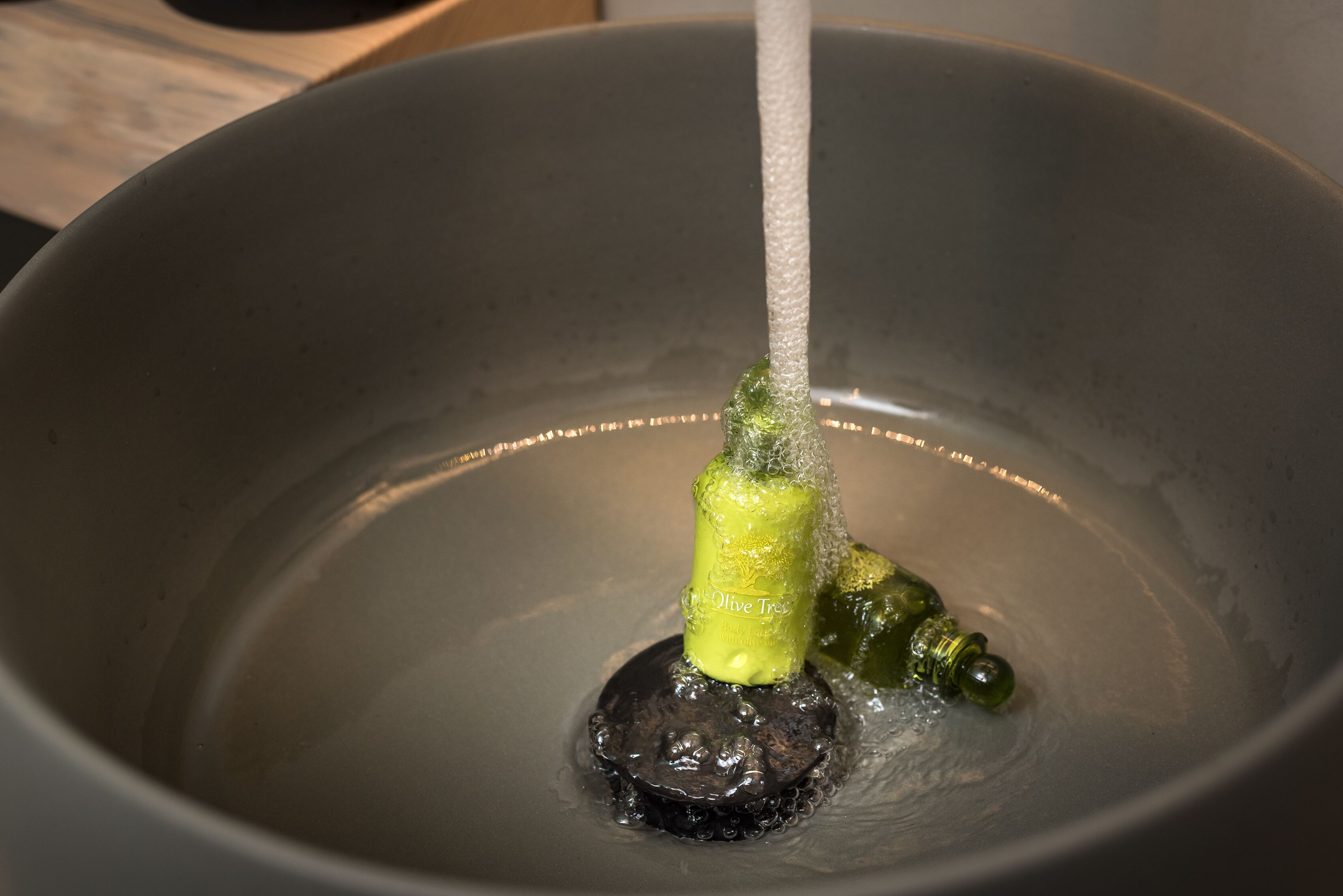 Un tazón con una botella verde etiquetada como 'Olive Tree' y un objeto oscuro sumergido en agua.
