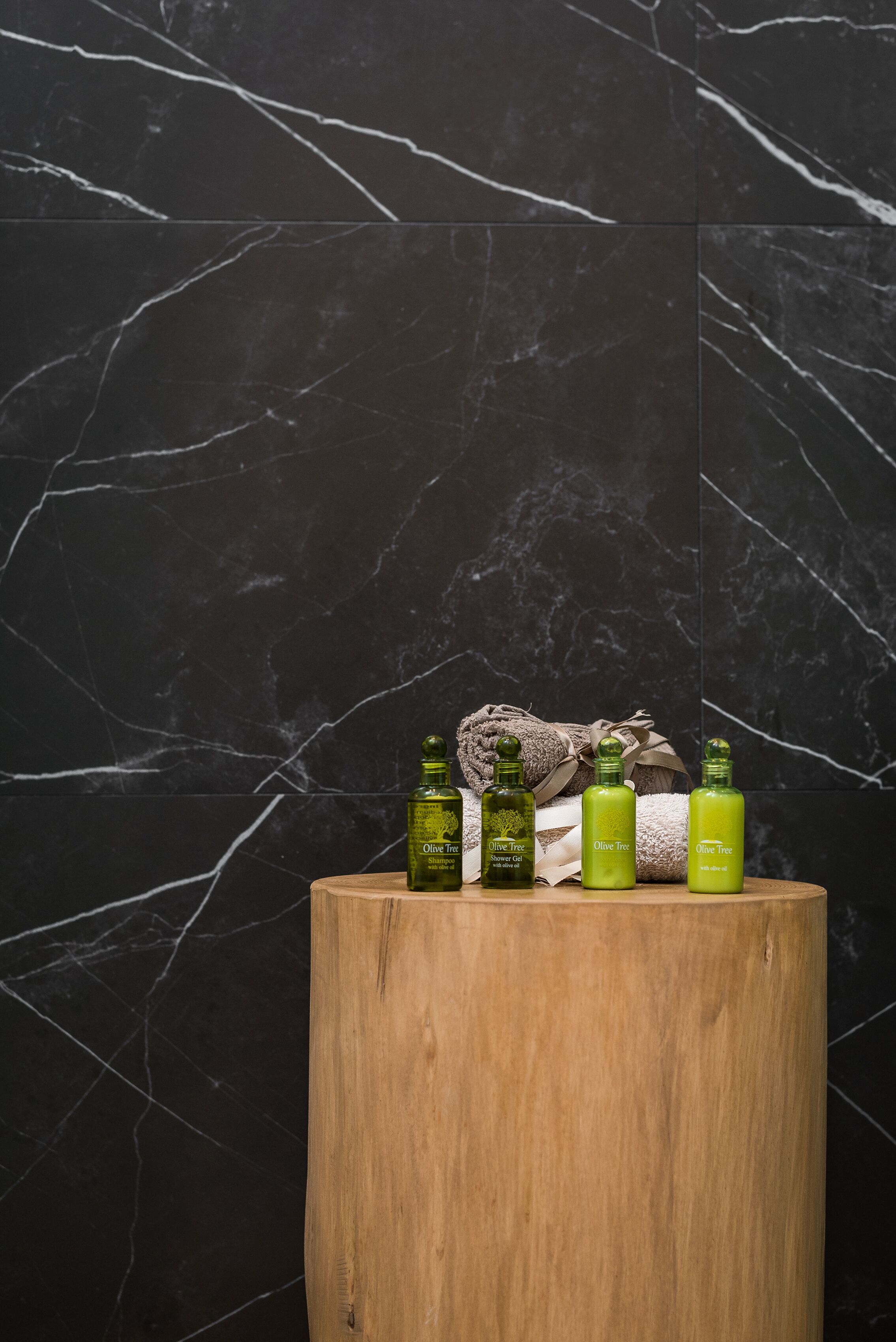 Een houten kruk met vier groene flessen, voorzien van het label 'Olive Tree', en een bruine handdoek op een zwarte marmeren oppervlakte.
