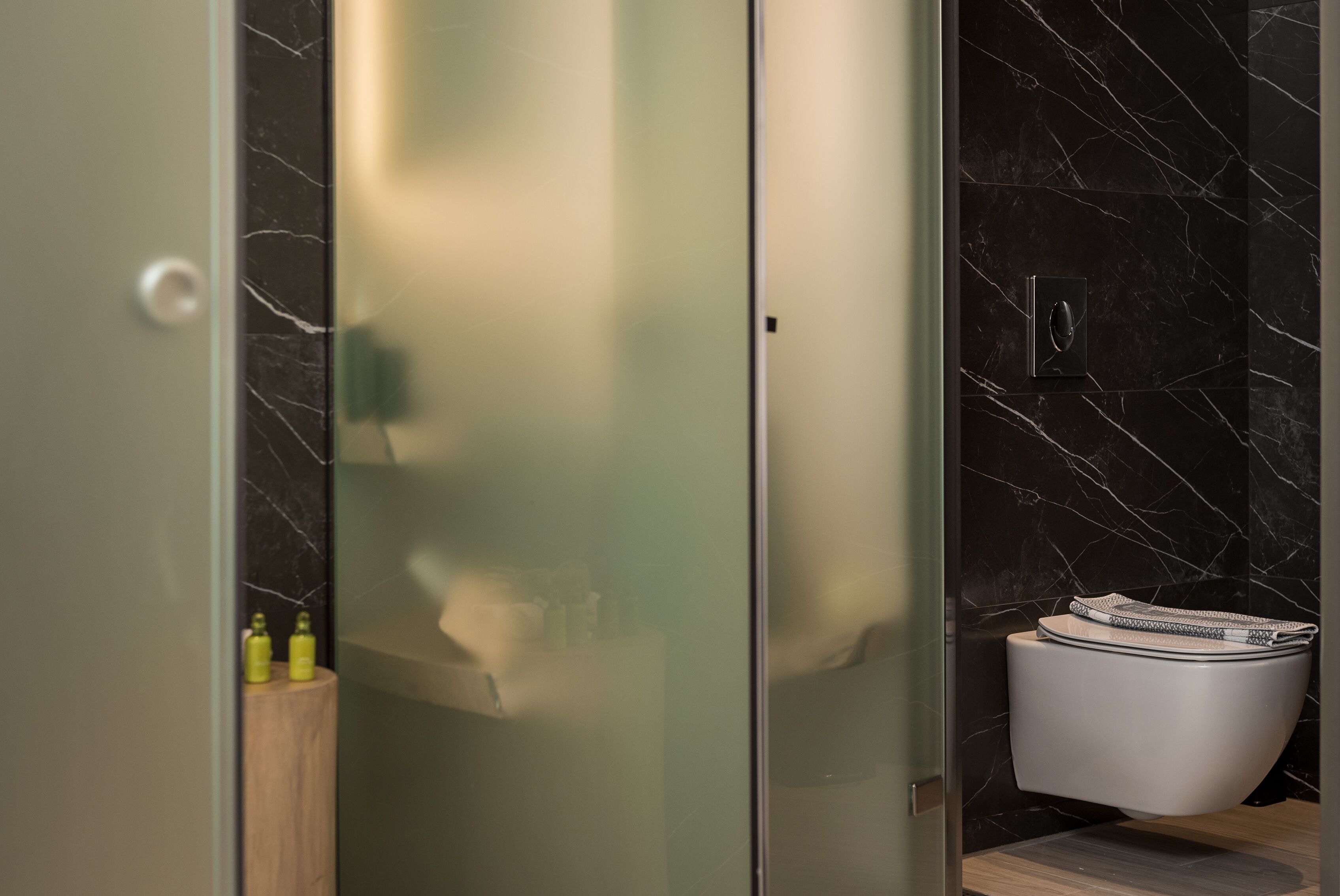 A modern bathroom with a glass shower enclosure, a marble-tiled wall, and a toilet with a magazine on top.
