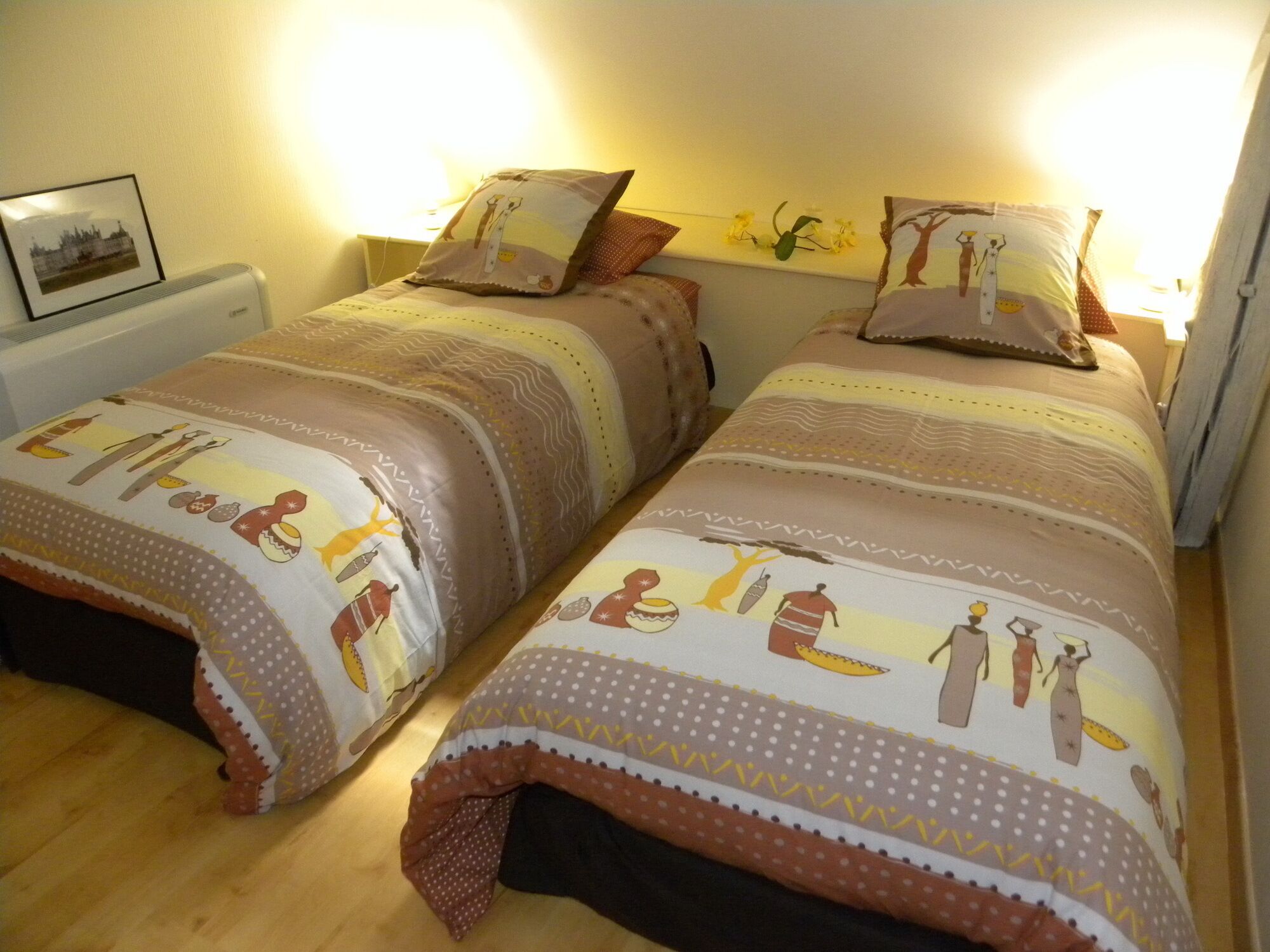 A bedroom with two beds, each with patterned bedding and pillows. There is a nightstand with a framed picture and a lamp between the beds.