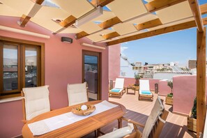 A sunlit patio with a wooden table and chairs, a view of the city, and a pink wall. - Evlimeni Suites (Chania)