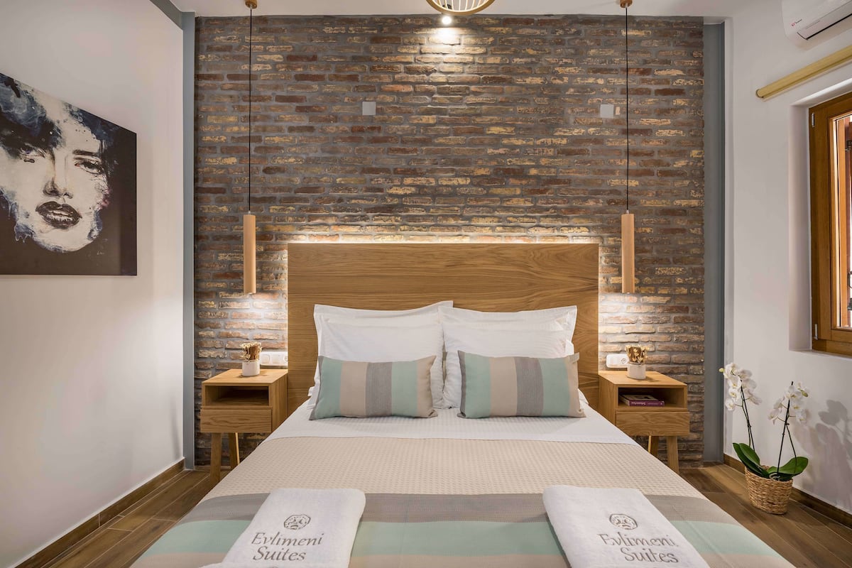 A hotel room with a brick wall, a wooden headboard, two bedside tables with lamps, a bed with striped pillows, and a framed picture on the wall.