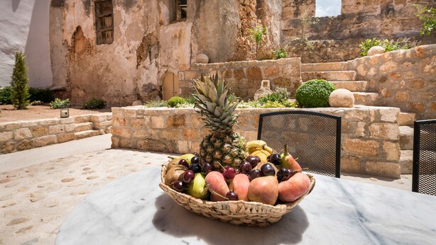 A fruit basket on a table in an outdoor setting with stone walls and a white building in the background. - Evlimeni Suites (Chania)