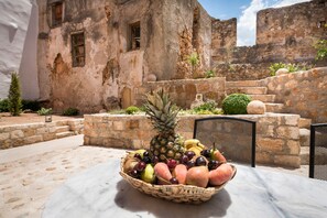 Un panier de fruits est posé sur une table dans un cadre extérieur, avec des murs en pierre et un bâtiment blanc en arrière-plan.