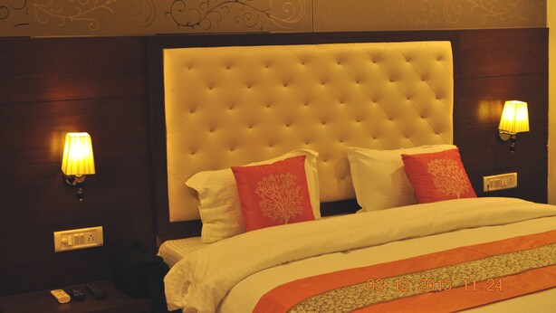 A hotel room with a tufted headboard, two bedside lamps, a bed with white and red bedding, and a nightstand with a remote control. - Hotel Grace (Gwalior)