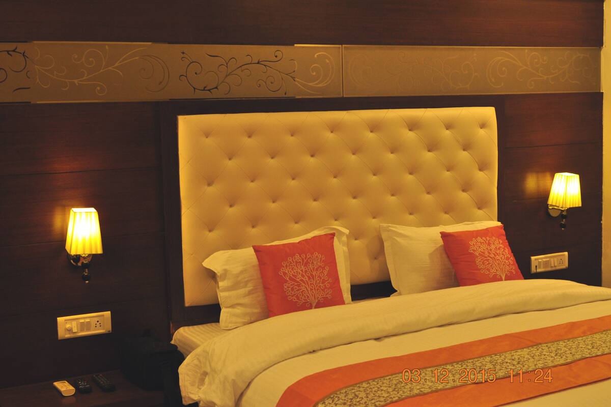 A hotel room with a tufted headboard, two bedside lamps, a bed with white and red bedding, and a nightstand with a remote control.