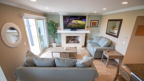 A living room with a grey sofa, a TV, a fireplace, and a dining area. - Oceanfront Boardwalk property, Newly remodeled Large patio, Steps to the sand. (Newport Beach)