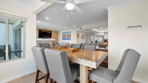 A modern dining area with a wooden table and grey upholstered chairs, a ceiling fan, recessed lighting, and a kitchen in the background. - Oceanfront Boardwalk property, Newly remodeled Large patio, Steps to the sand. (Newport Beach)