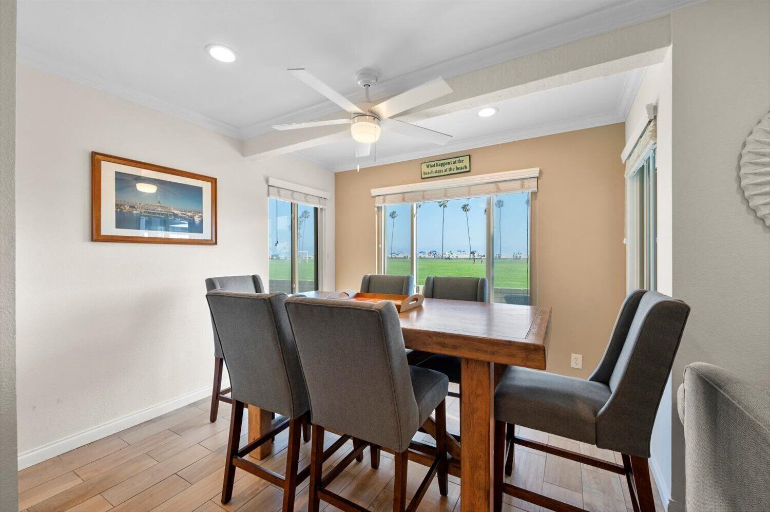 A dining area with a table and chairs, a ceiling fan, a framed picture, and a view of the beach through large windows.