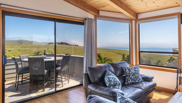 A room with a leather sofa, a dining table with chairs, and large windows offering a view of a grassy landscape and the sea. - Beautiful South Harbor Home with Great Ocean Views  Point View House (Bodega Bay)