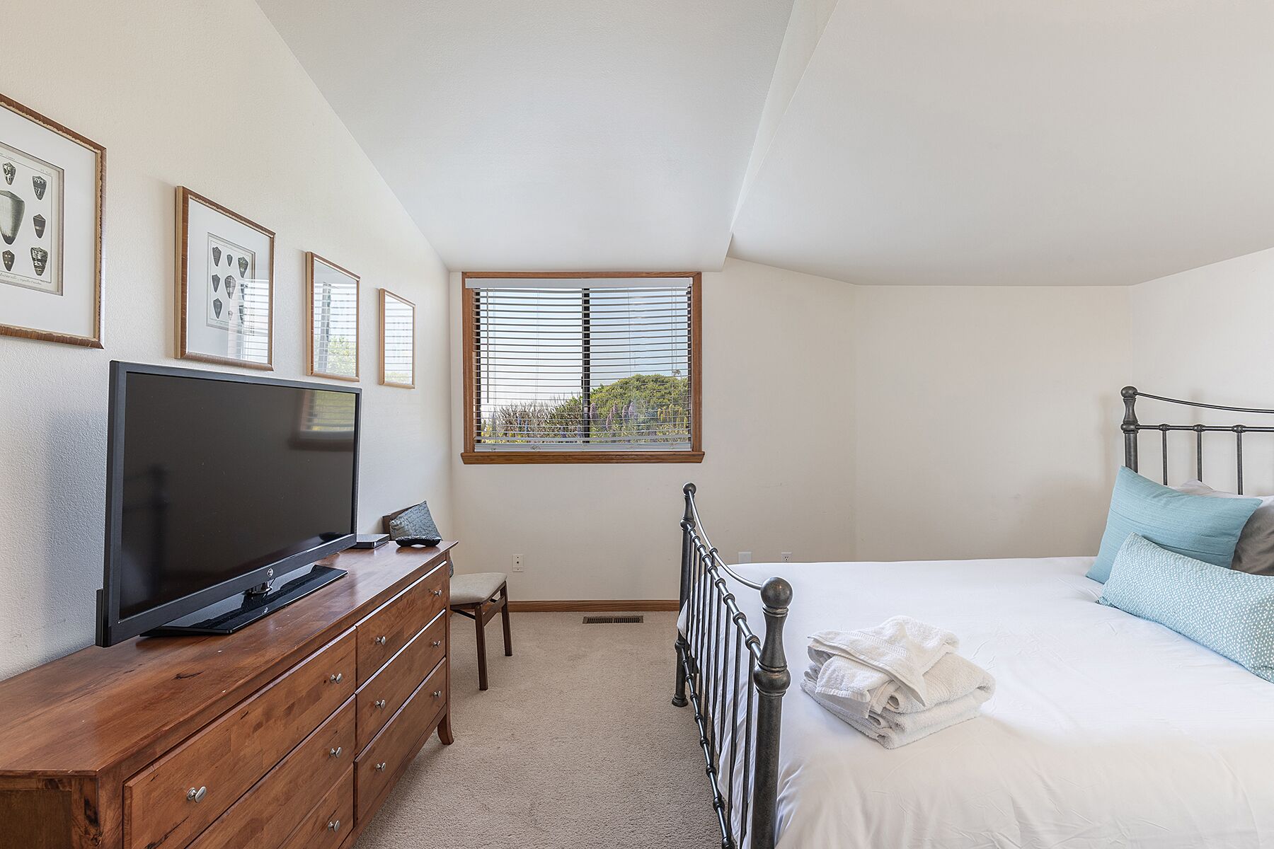 A bedroom with a bed, a TV on a wooden dresser, a window with blinds, and framed pictures on the wall.