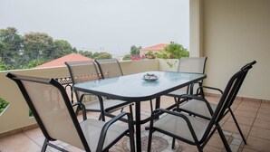 Un balcón con una mesa y sillas de vidrio, piso de baldosas y vista a edificios y árboles al fondo.