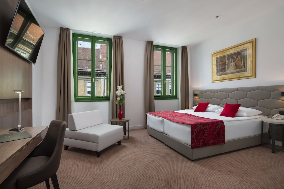 Ein Hotelzimmer mit Bett, Schreibtisch, Stuhl, einem kleinen Tisch mit einer Vase und einem Gemälde an der Wand.