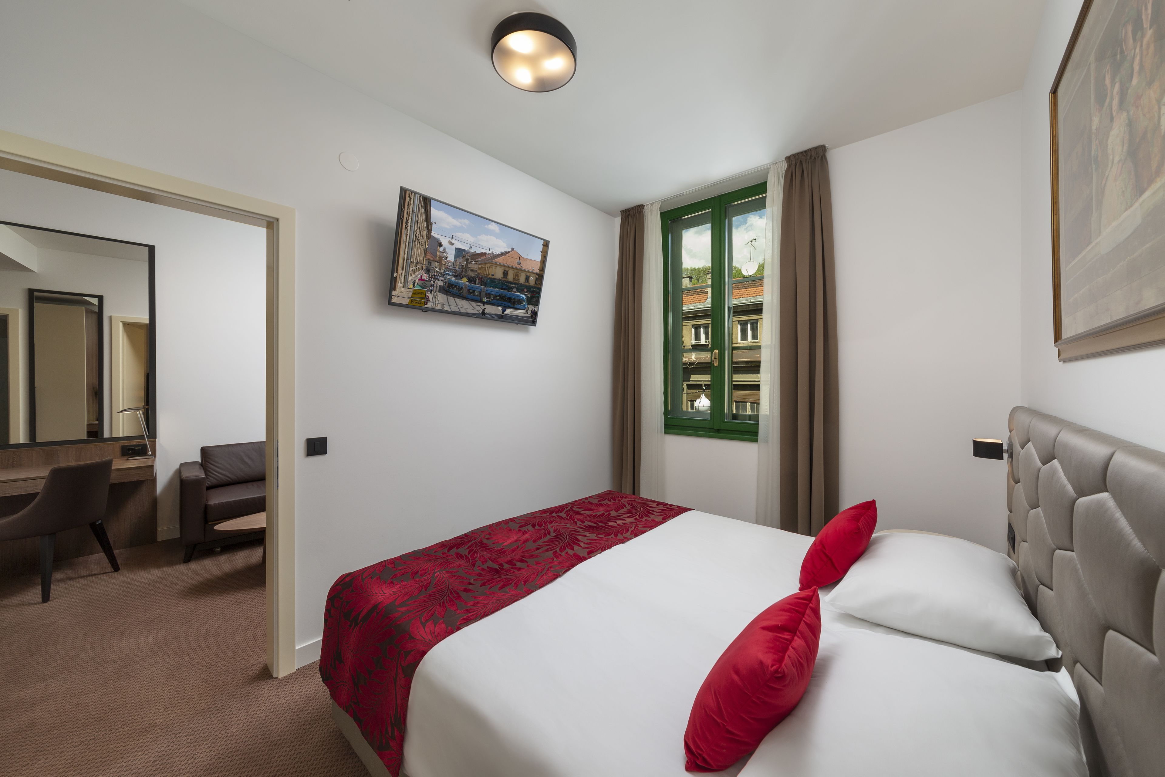 A hotel room with a bed, a desk, a chair, a painting, and a window with curtains.