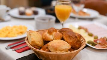 Une table mise pour le petit-déjeuner, avec un panier de petits pains, un verre de jus d’orange et des assiettes remplies de nourriture.