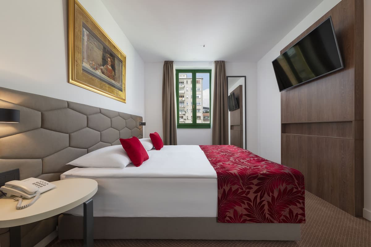Ein Hotelzimmer mit einem Bett, einem Nachttisch mit Telefon, einem an der Wand montierten Fernseher und einem Fenster mit Vorhängen.