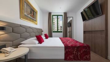 Une chambre d’hôtel avec un lit, une table de chevet avec un téléphone, un téléviseur fixé au mur et une fenêtre avec des rideaux.