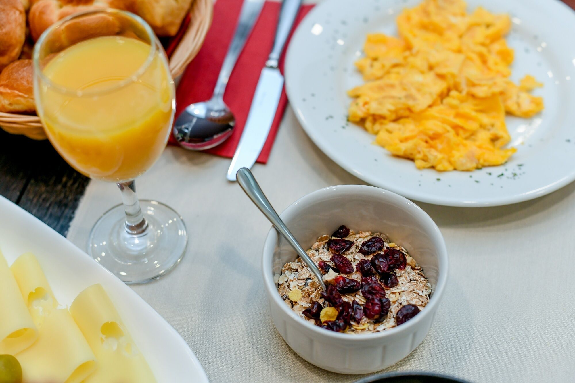 A breakfast setting with a glass of orange juice, scrambled eggs, a bowl of cereal with raisins, and a plate of cheese slices.