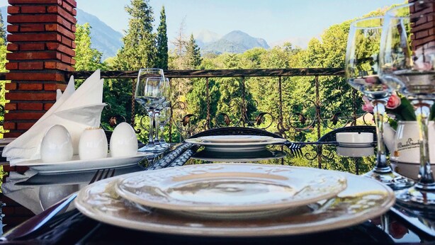 An outdoor dining table with plates, glasses, and a basket, set against a backdrop of trees and mountains.
