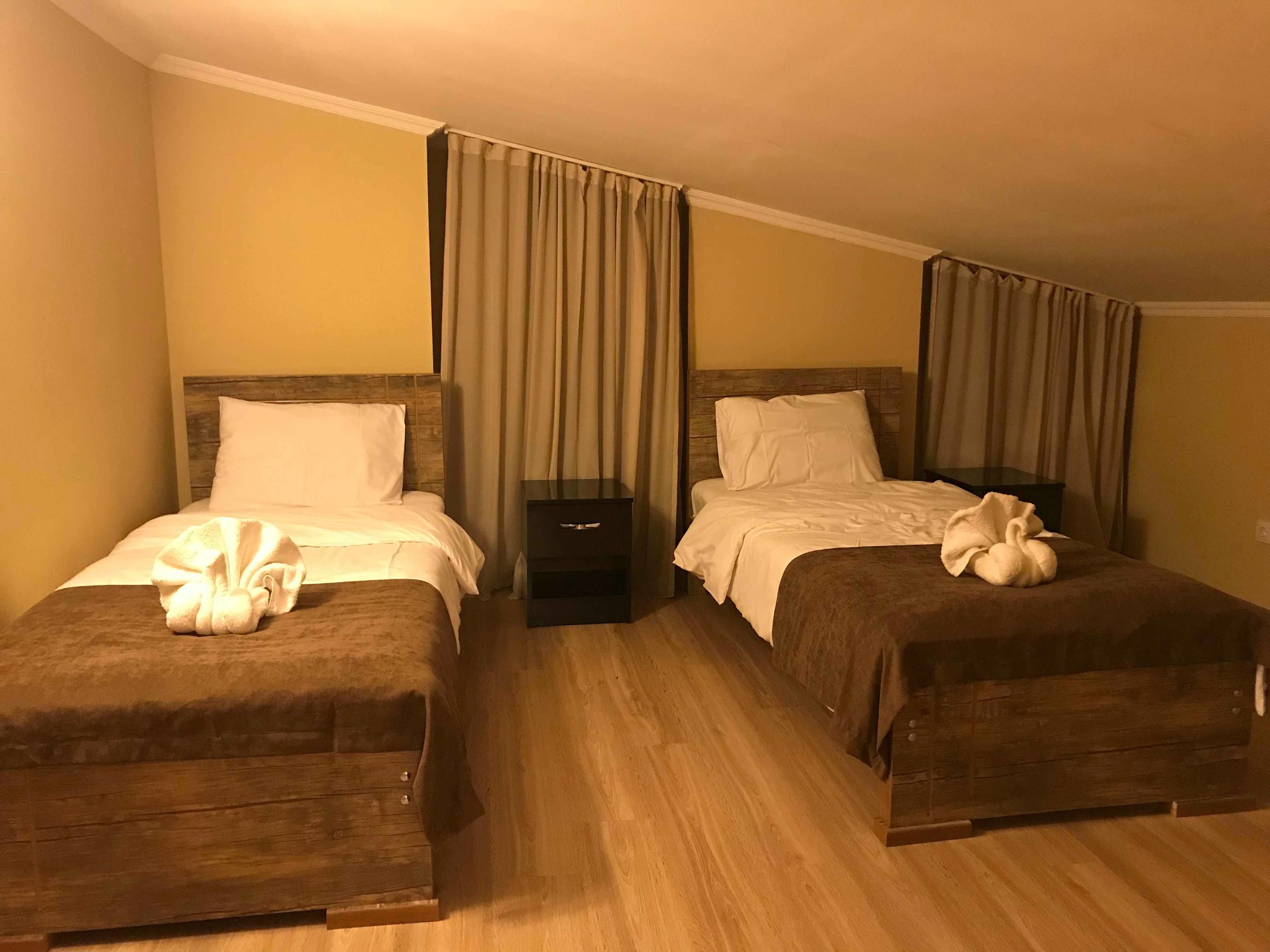 Habitación de hotel con dos camas, cada una con cabecero de madera, ropa blanca y una toalla sobre las camas. Hay una mesita de noche pequeña entre las camas.