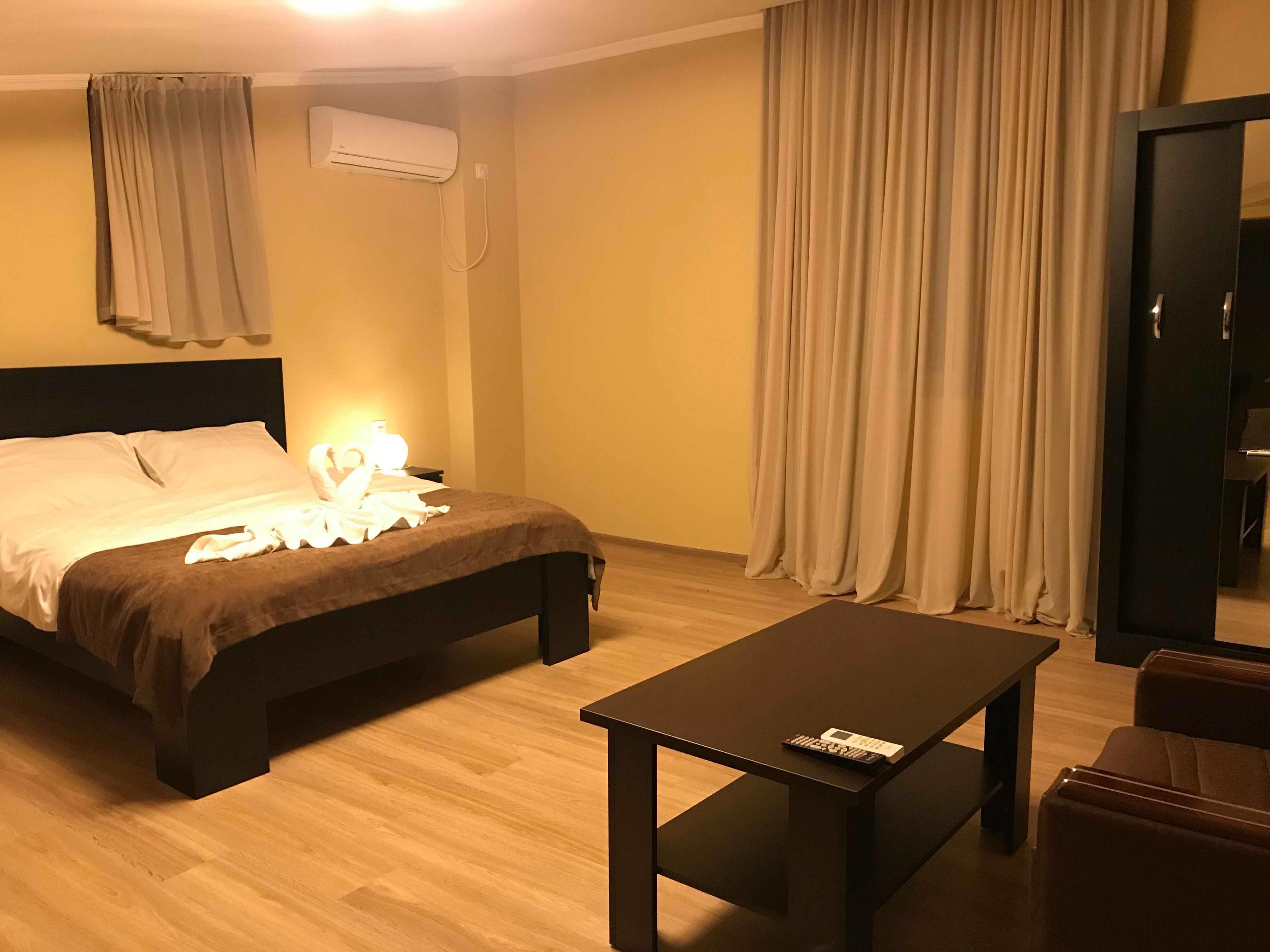 A hotel room with a bed, a bedside table, a chair, a desk, an air conditioning unit, and curtains.