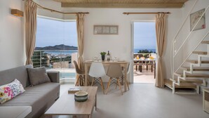 A modern living room with a sofa, dining table, and chairs, a staircase, and large windows with a view of the sea. - Pantanassa Villas (Chania)
