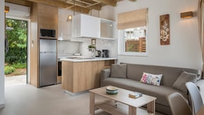 A modern kitchen with a built-in microwave, refrigerator, and sink. There is a wooden island with a sink and a coffee maker. A beige sofa and a small table with a bowl and book are in the living area. - Pantanassa Villas (Chania)
