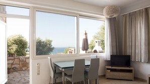 A room with a dining table, chairs, a TV on a wooden stand, and a view of the sea through large windows. - Pantanassa Villas (Chania)