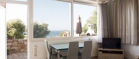 A room with a dining table, chairs, a TV on a wooden stand, and a view of the sea through large windows.