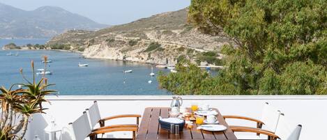 Una mesa de madera preparada para el desayuno con sillas blancas, con vistas a una zona costera con barcos y colinas.