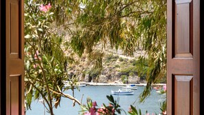 An open wooden door framing a view of a calm body of water with boats, surrounded by greenery and a rocky shoreline. - Villa Notos (Milos)