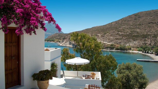 A white building with a wooden door, a balcony with a white umbrella, and a view of a blue sea and hills. - Villa Notos (Milos)