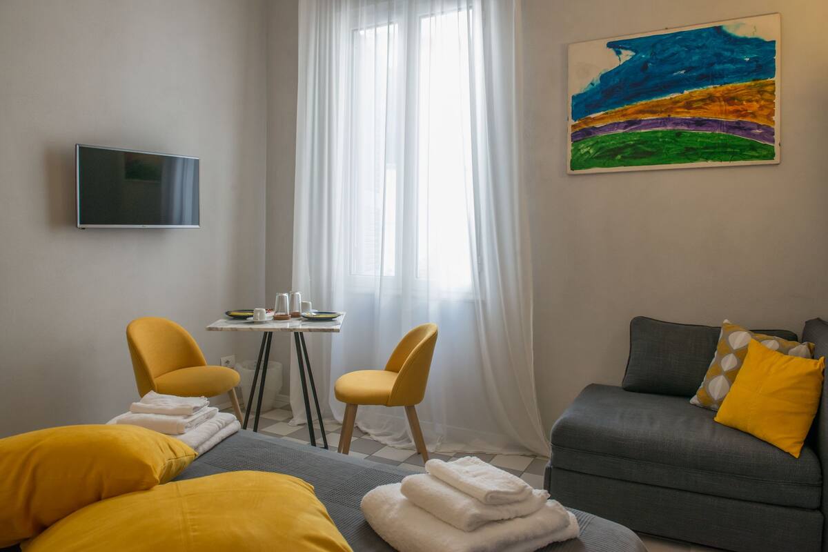 A hotel room with a bed, two yellow chairs, a grey sofa, a TV on the wall, and a painting.