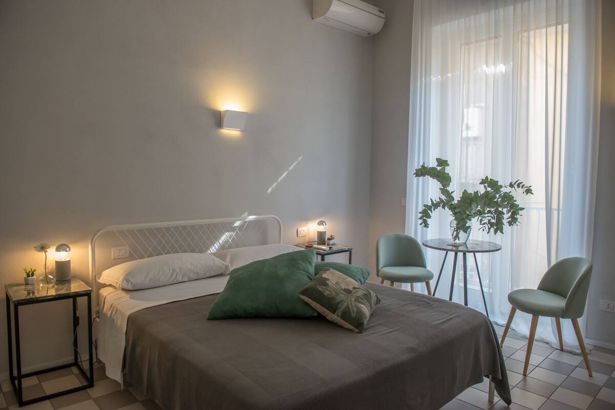 A modern bedroom with a bed, bedside tables, a small round table, two chairs, and a potted plant.