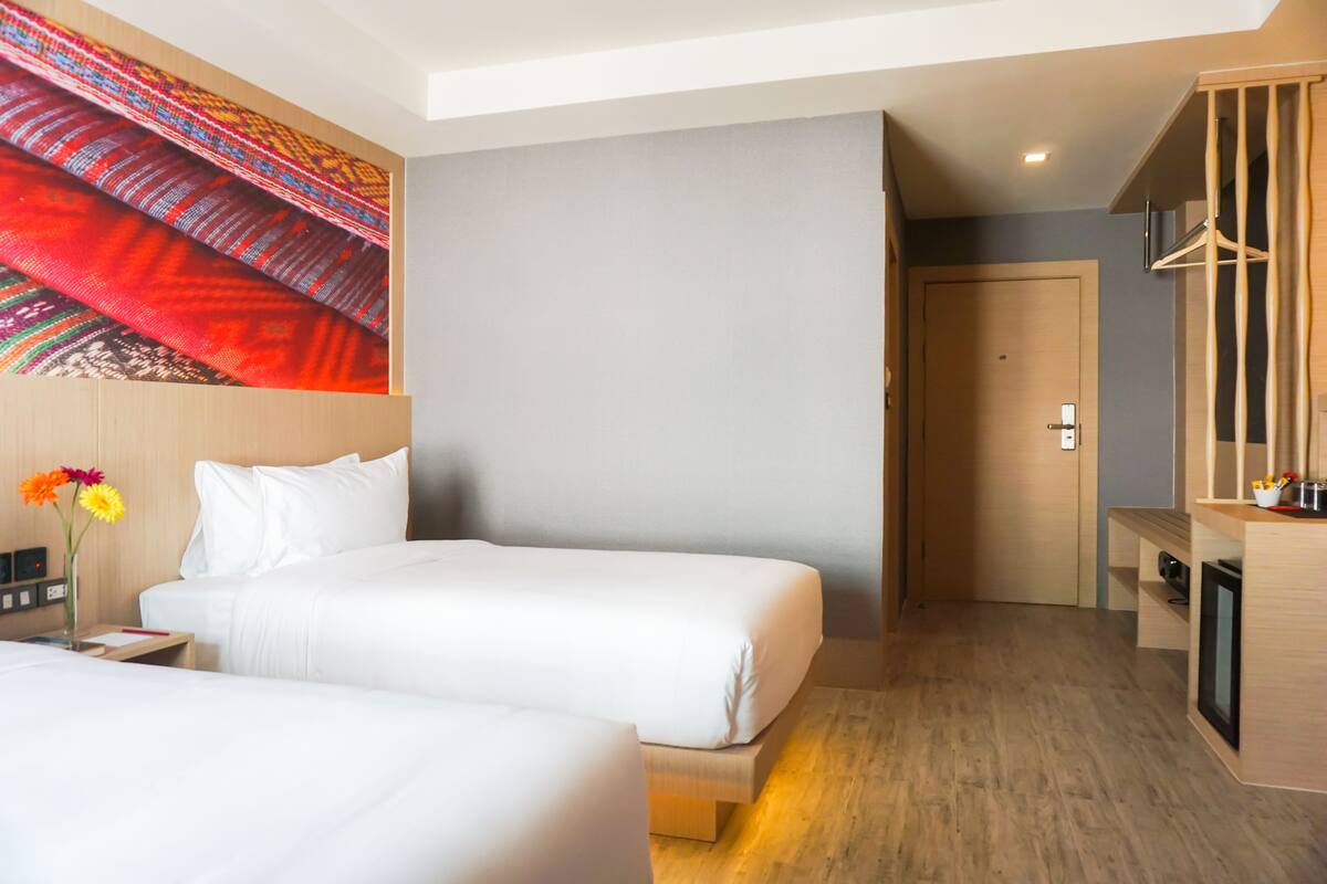 Une chambre d’hôtel avec deux lits, un plancher en bois, un mur orné d’une tapisserie colorée, une porte et un petit bureau sur lequel est posé un vase de fleurs.