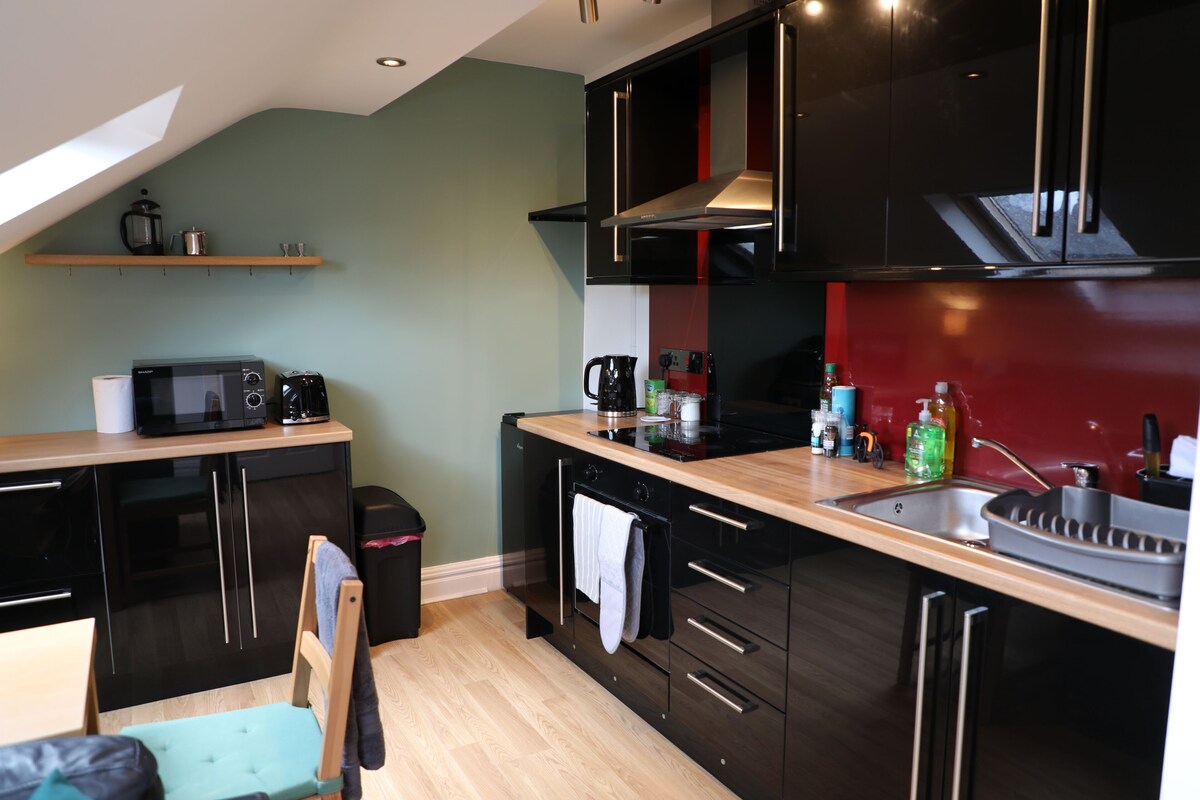 A modern kitchen with black cabinets, a wooden countertop, and a red backsplash. There is a microwave, toaster, and coffee maker on the counter. A chair is visible in the foreground.