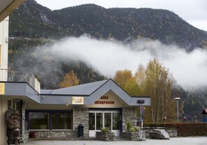 Un bâtiment appelé « KRO RESEPSION », doté d’une façade en pierre, de grandes fenêtres et ayant une chaîne de montagnes en toile de fond.