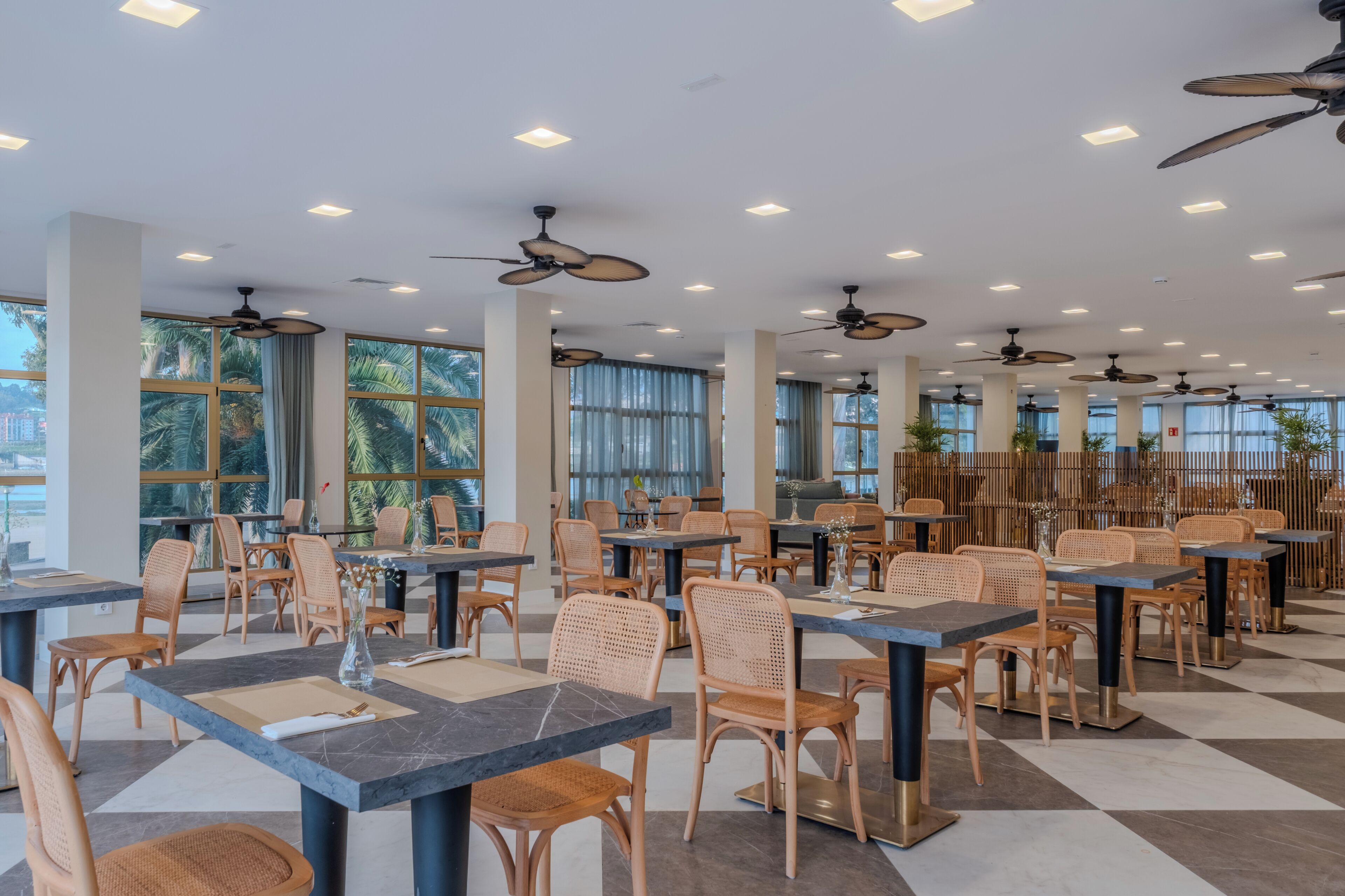 Un comedor interior espacioso con techos altos, ventiladores de techo y ventanas amplias. Las mesas están dispuestas en grupos con sillas de mimbre.