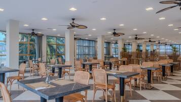 Un comedor interior espacioso con techos altos, ventiladores de techo y ventanas amplias. Las mesas están dispuestas en grupos con sillas de mimbre.