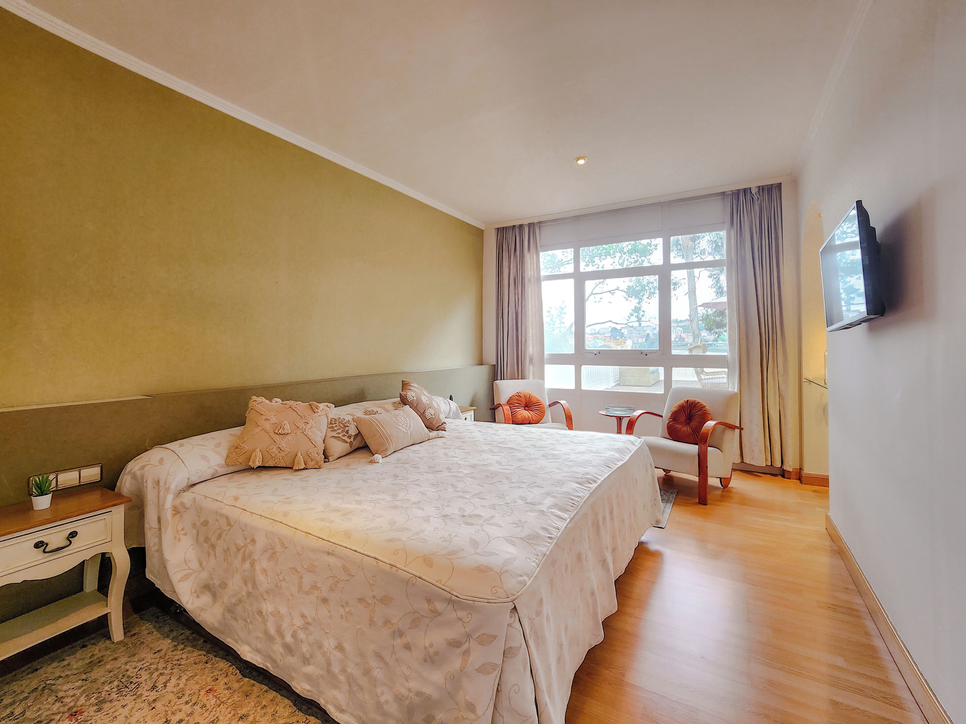 A bedroom with a large bed, bedside tables, a TV mounted on the wall, and a window with curtains.