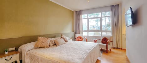 A bedroom with a large bed, bedside tables, a TV mounted on the wall, and a window with curtains.