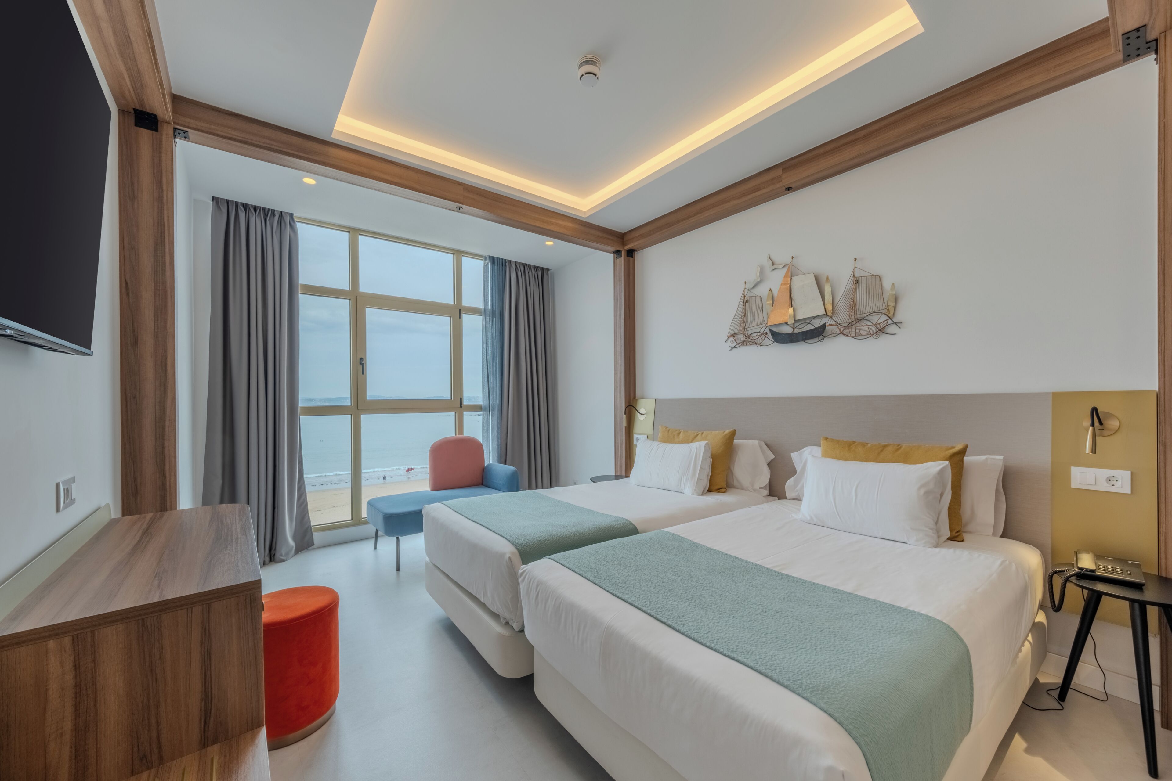 Habitación de hotel con dos camas, un escritorio, un otoman rojo, una silla, un televisor y un ventanal con cortinas.