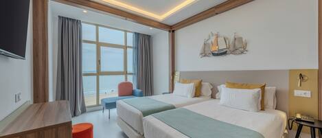 A hotel room with two beds, a desk, a red ottoman, a chair, a TV, and a large window with curtains.