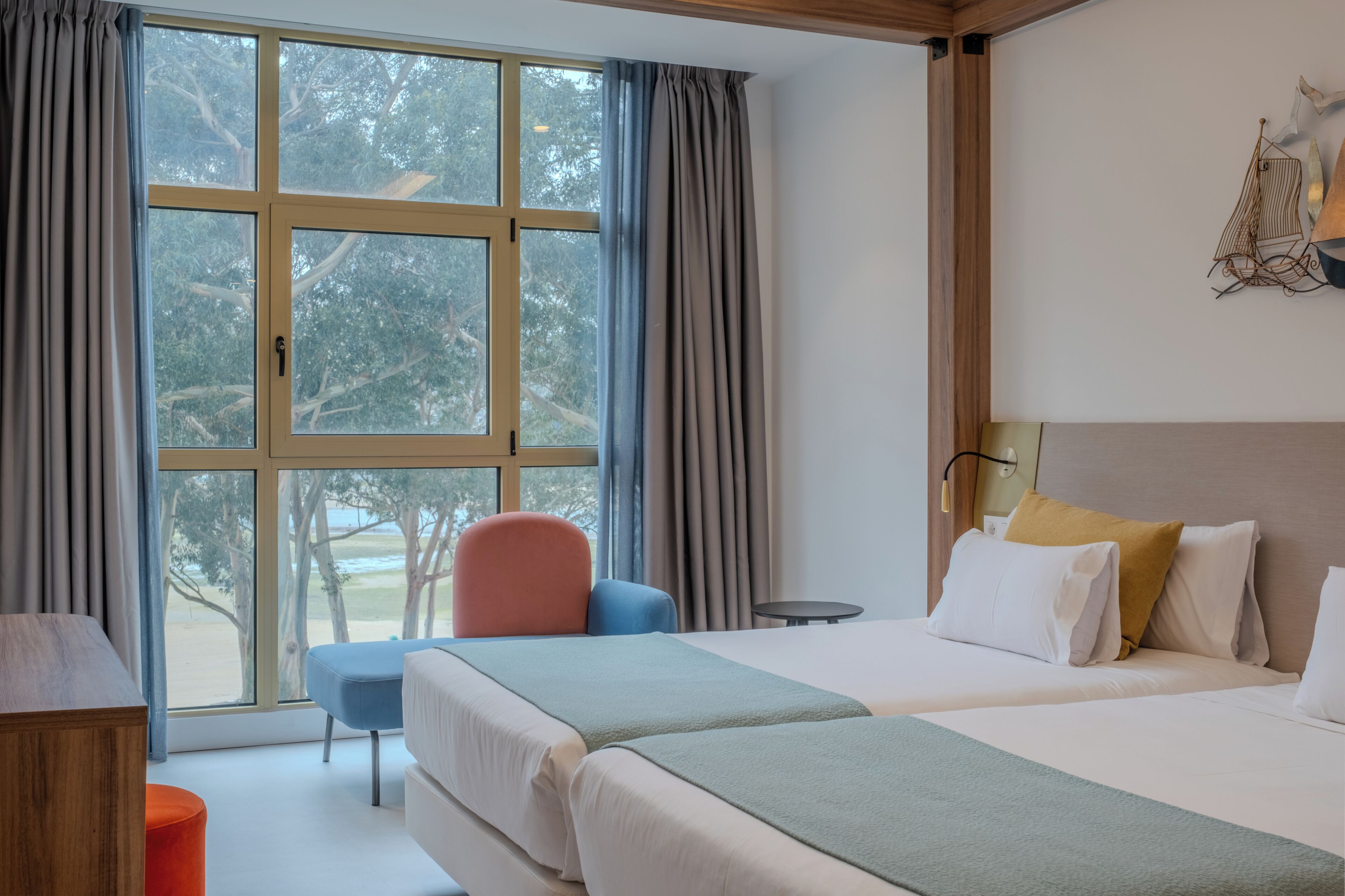 A hotel room with a large window, a bed, a chair, a small table, and a lamp.